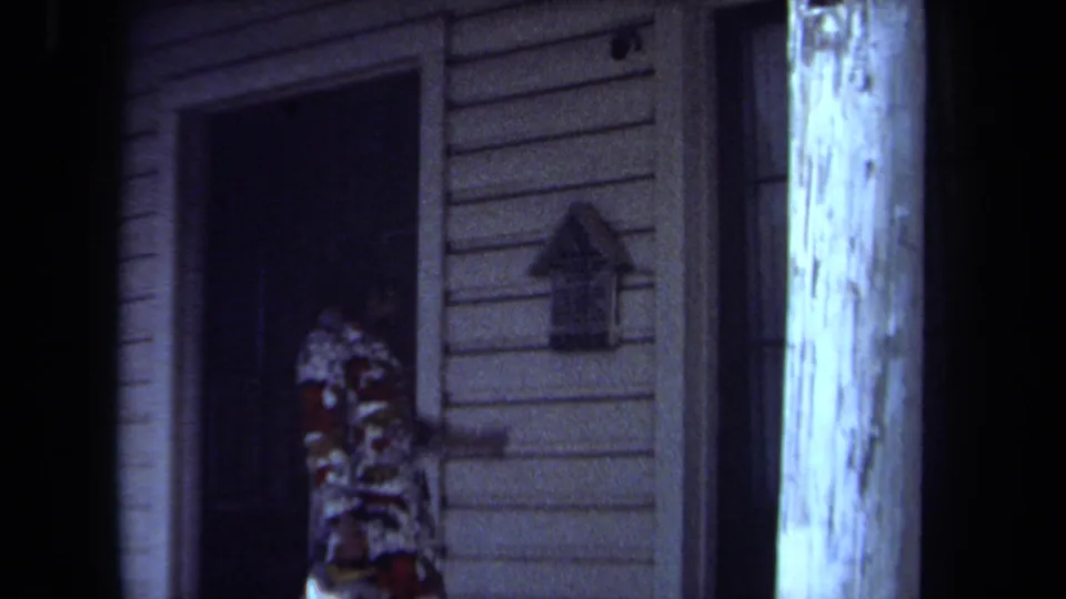 1971 Maine home-movie frame showing the woman partly shadowed near the doorway, with a wall mailbox and white porch post visible.