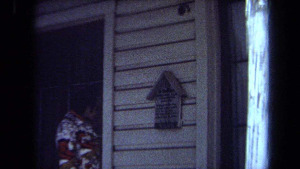 1971 Maine 8mm frame showing a partially seen adult at a dark doorway and a small house-shaped sign on a clapboard exterior.