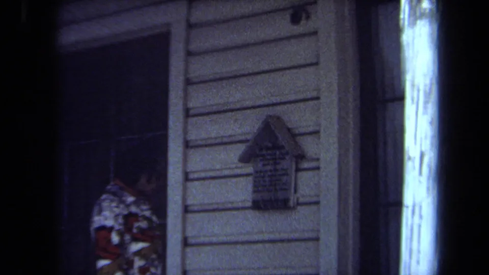 1971 Maine 8mm frame showing a partially seen adult at a dark doorway and a small house-shaped sign on a clapboard exterior.