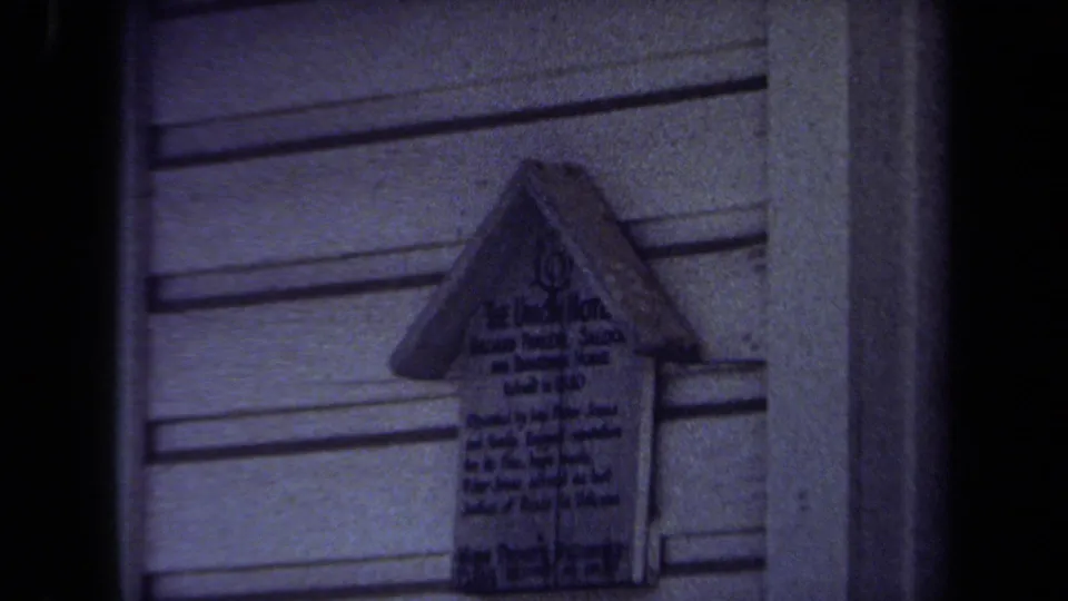 1971 Maine 8mm frame centered on a small house-shaped wooden marker with dark handwritten text on a clapboard wall.