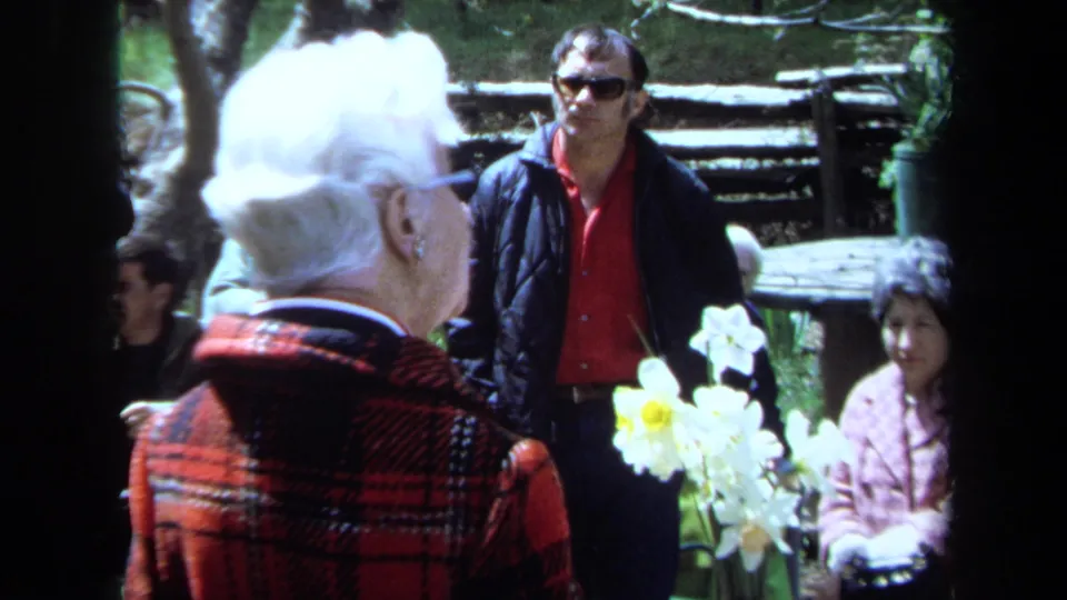 An elderly woman in a red plaid jacket stands with white flowers before attendees at an outdoor 1971 Maine memorial gathering.