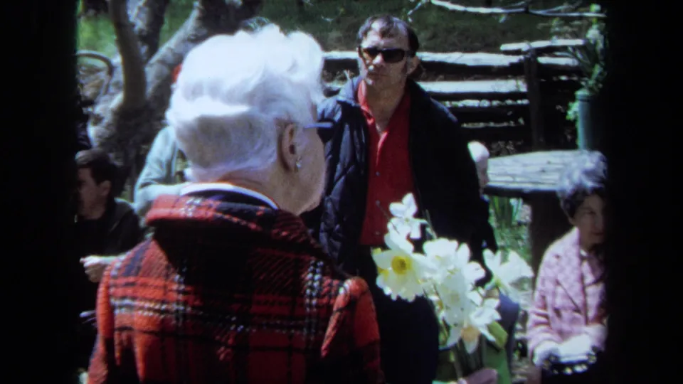 The frame shows the elderly woman from behind with flowers, a man in sunglasses, and nearby attendees during the Maine memorial scene.