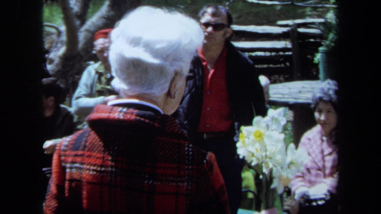 In 1971 Maine home-movie footage, an older woman seen from behind listens with other garden visitors near white daffodils.