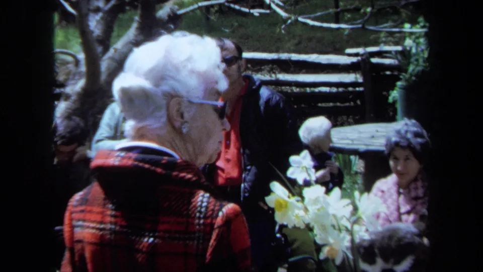 The older woman in red plaid is shown in profile during the Maine garden visit, with white daffodils and other guests nearby.