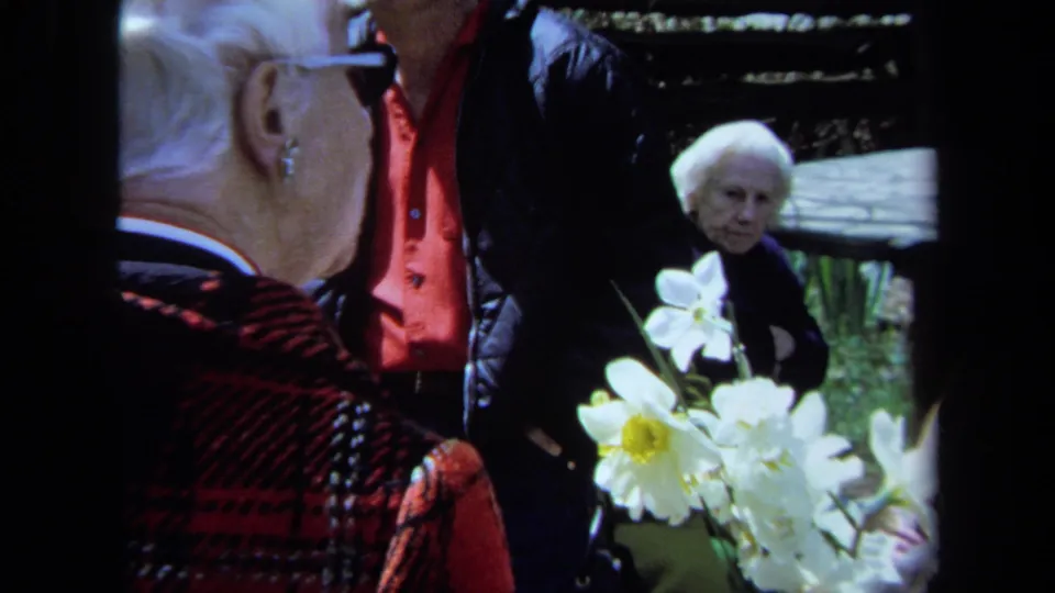 A close 8mm frame shows an older woman in red plaid near white daffodils, with another older visitor and a man behind her.