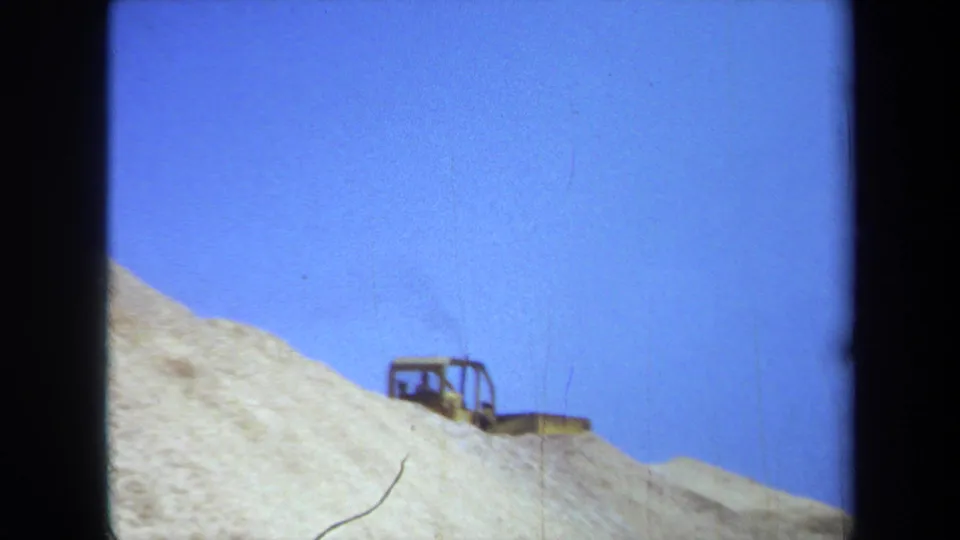 A distant construction vehicle sits on a sandy mound under clear blue sky in 1971 Maine footage.