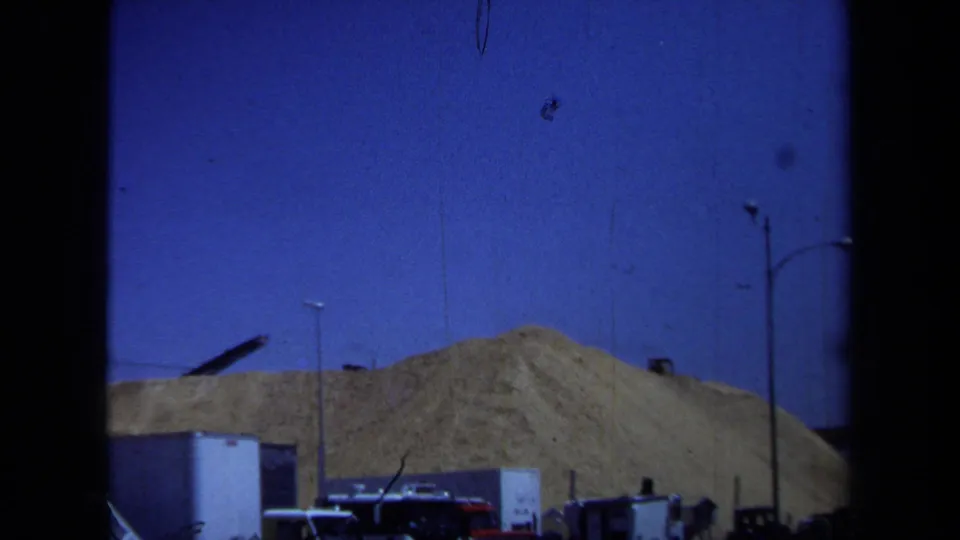 A wider construction view shows sandy mounds, parked cars, utility poles, and a few distant indistinct figures or equipment shapes.