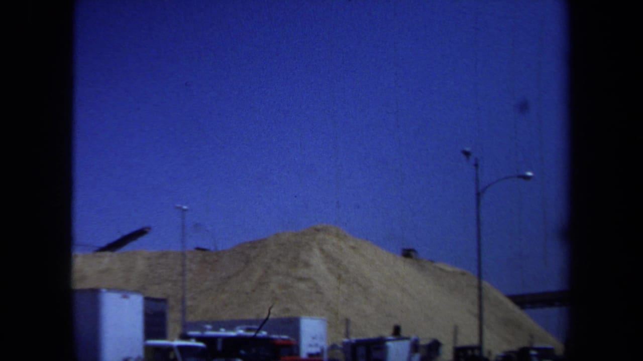 1971 Maine 8mm frame showing a large aggregate pile, parked trucks, a streetlight, and industrial yard structures under a deep blue sky.