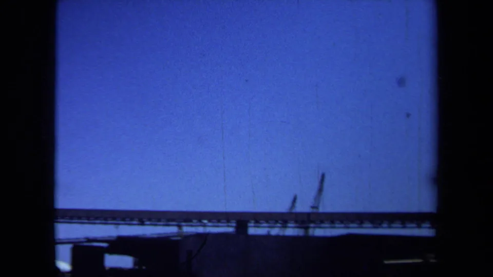 1971 Maine 8mm frame showing a long conveyor line, low industrial roofline, and distant crane-like booms beneath an open blue sky.