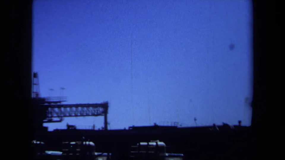 Maine 1971 8mm frame with industrial trusswork, low vehicles, and open blue sky in a static home-movie view.