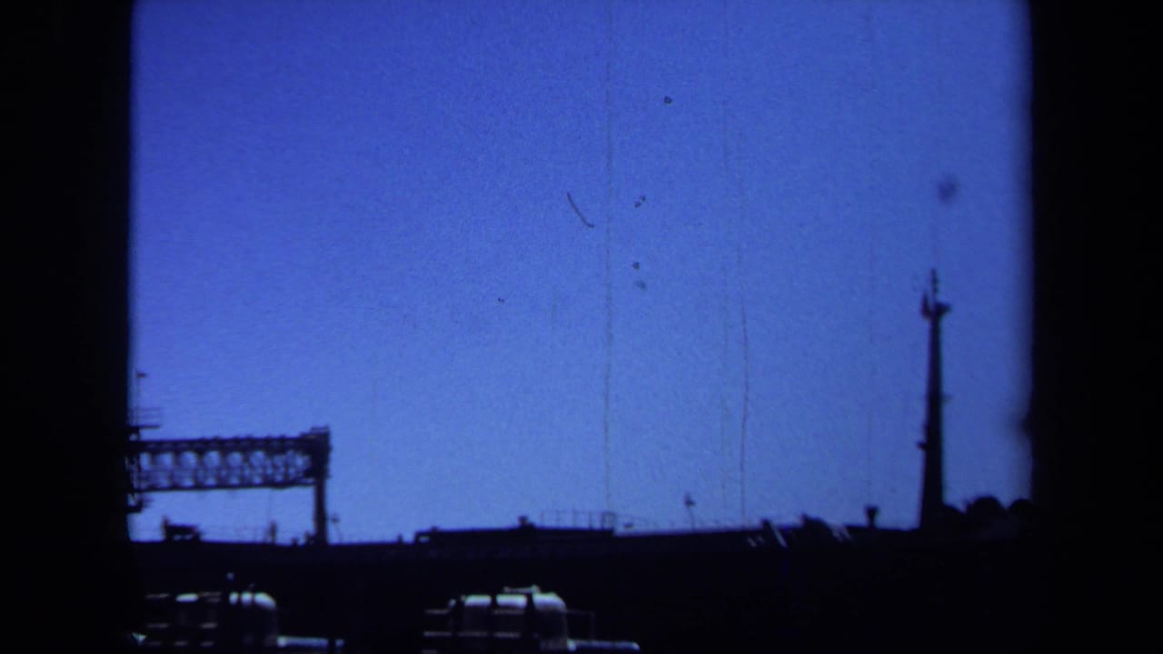 1971 Maine 8mm frame showing a far-off steel bridge or industrial span in silhouette beneath a blue sky.