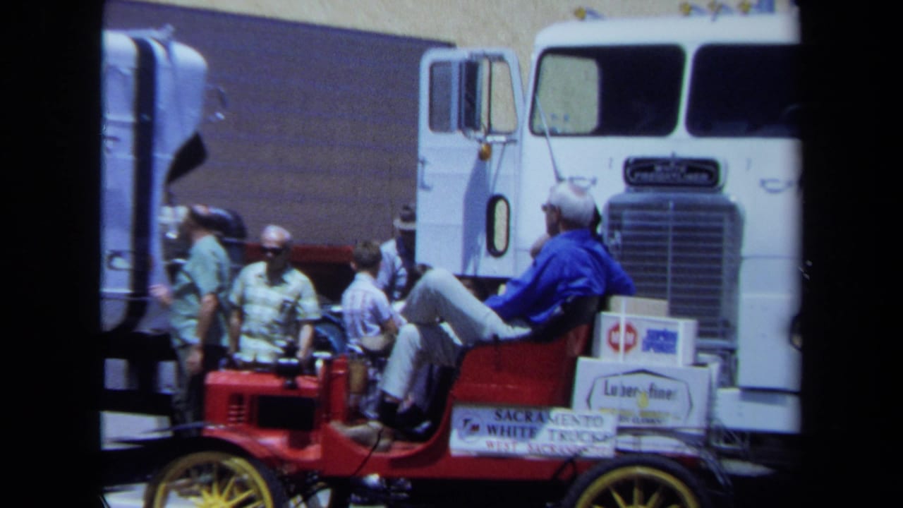 Man sits in a red antique car beside a white semi-truck as other men stand nearby.