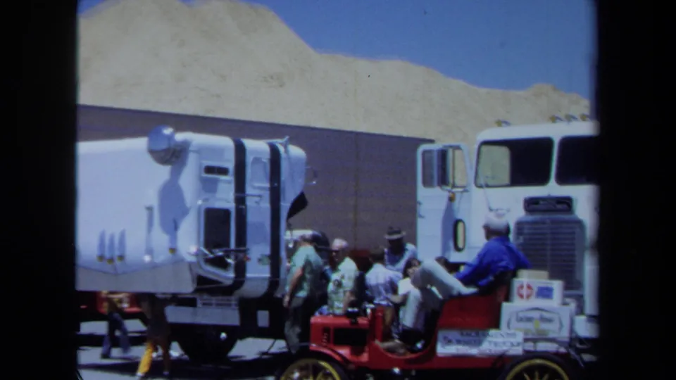 1971 Maine frame showing a red antique car with yellow wheels near modern white semi-trucks and men in casual clothing.