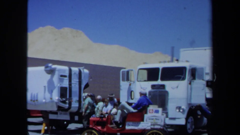 Wide 1971 Maine 8mm view of men around a red antique car parked by white semi-trucks with sand-colored mounds behind.