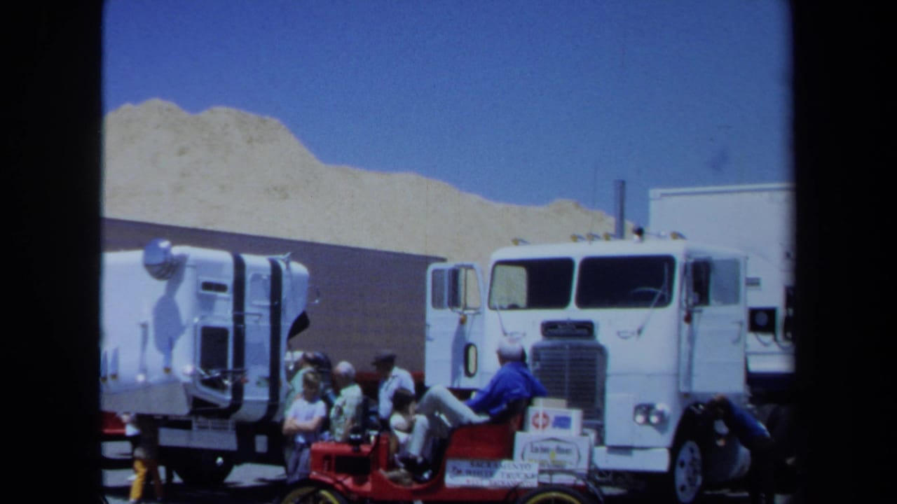 White semi-truck beside a trailer with several men gathered near a small red tractor.