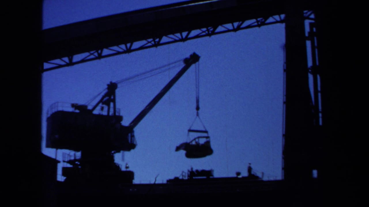 A large crane appears in silhouette beneath an overhead structure, lifting equipment against a deep blue sky in the Maine worksite footage.