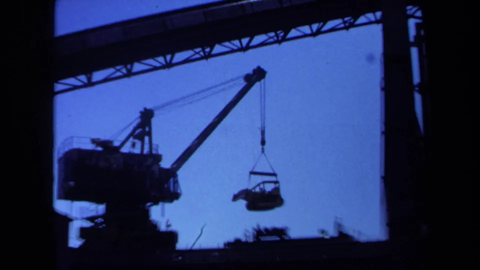 The crane boom and hook are silhouetted while a suspended piece of machinery hangs over the work area in this 1971 Maine 8mm frame.