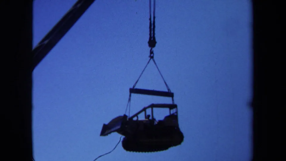 A closer 8mm view shows heavy machinery hanging from crane rigging against the blue sky, with the object mostly in silhouette.
