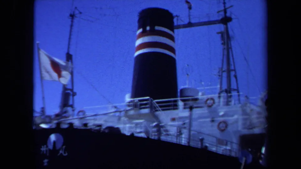 Dockside 8mm view of the Japanese cargo ship in Maine, with flag, funnel bands, and upper deck structures.