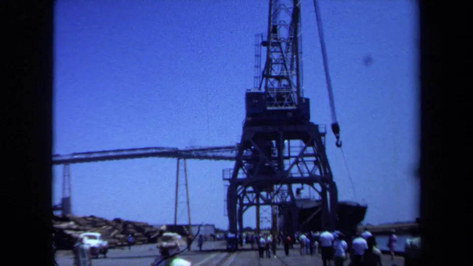 1971 Maine port view with a tall industrial crane, pier activity, parked cars, and visitors gathered near the waterfront.