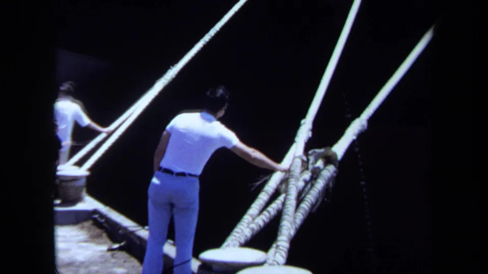 1971 Maine 8mm frame showing men near boat rigging, with one man holding thick ropes beside a dark waterfront or vessel area.