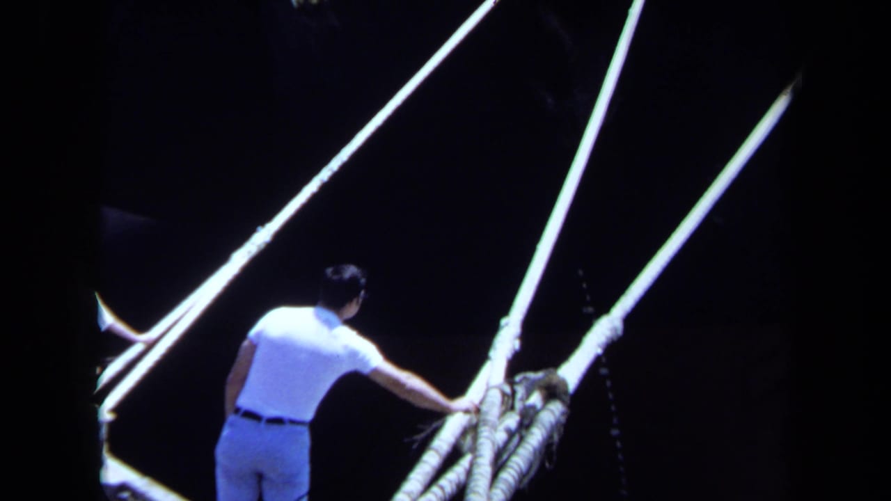 In Maine 1971 home-movie footage, a man on a dock reaches toward heavy mooring lines beside a dark Japanese vessel hull.