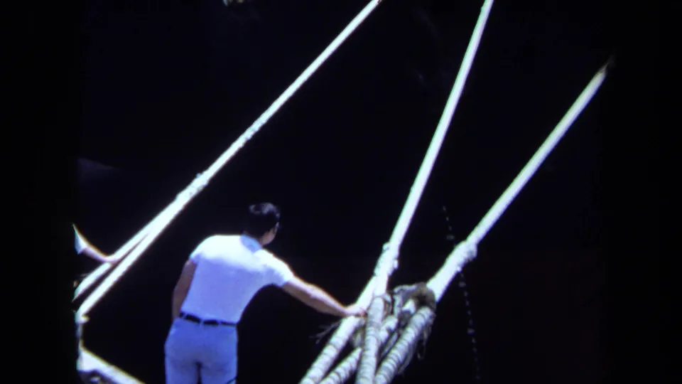 In Maine 1971 home-movie footage, a man on a dock reaches toward heavy mooring lines beside a dark Japanese vessel hull.
