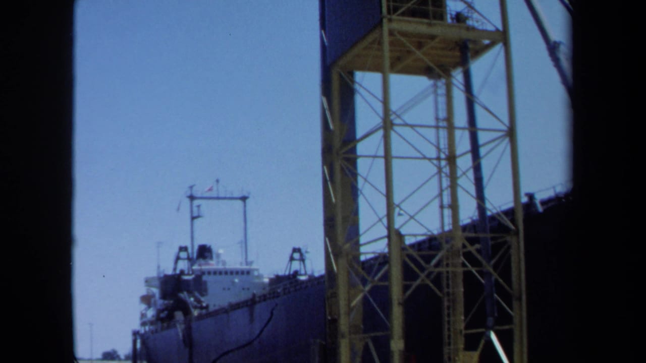 1971 Maine home-movie frame with a cargo ship at dock, a tall yellow truss structure, and a crane or boom line against clear sky.