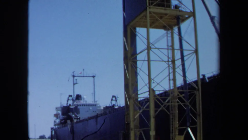 1971 Maine home-movie frame with a cargo ship at dock, a tall yellow truss structure, and a crane or boom line against clear sky.
