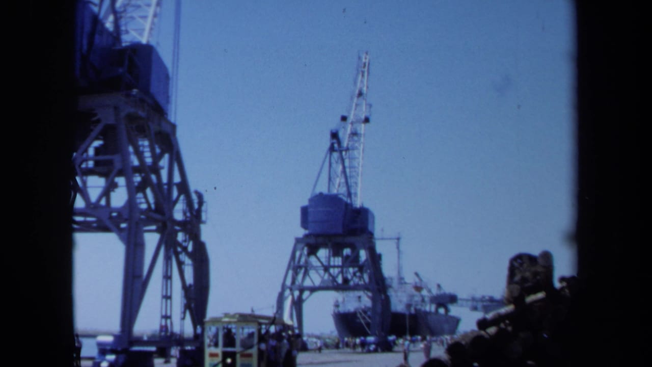 1971 Maine home-movie frame showing industrial port cranes, a pier roadway, a small vehicle, and a ship at the dock.