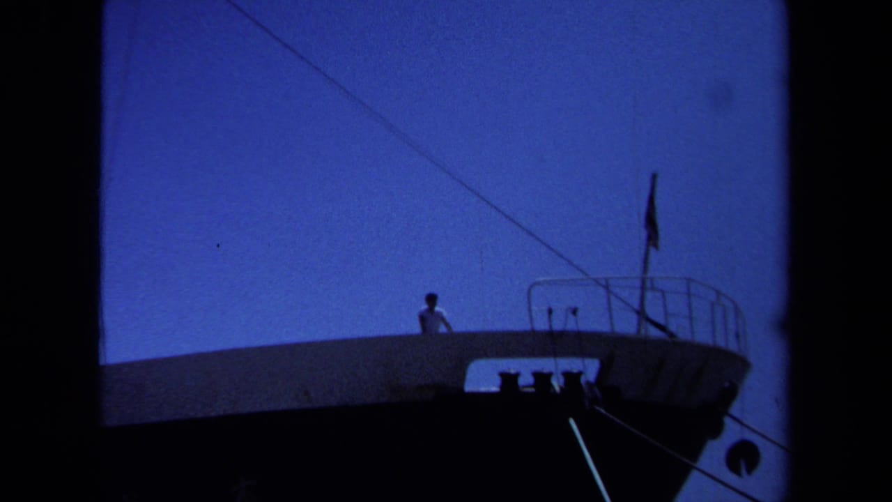 1971 Maine waterfront frame with a distant man in a white shirt on a vessel deck near railings and rigging lines.