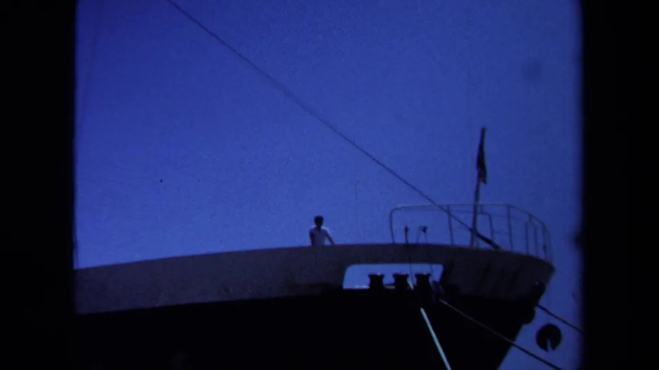 1971 Maine waterfront frame with a distant man in a white shirt on a vessel deck near railings and rigging lines.