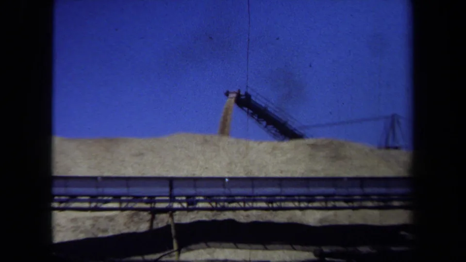 1971 Maine 8mm frame showing a grain mound, distant conveyor, and industrial rails or pipes from a handheld vantage point.
