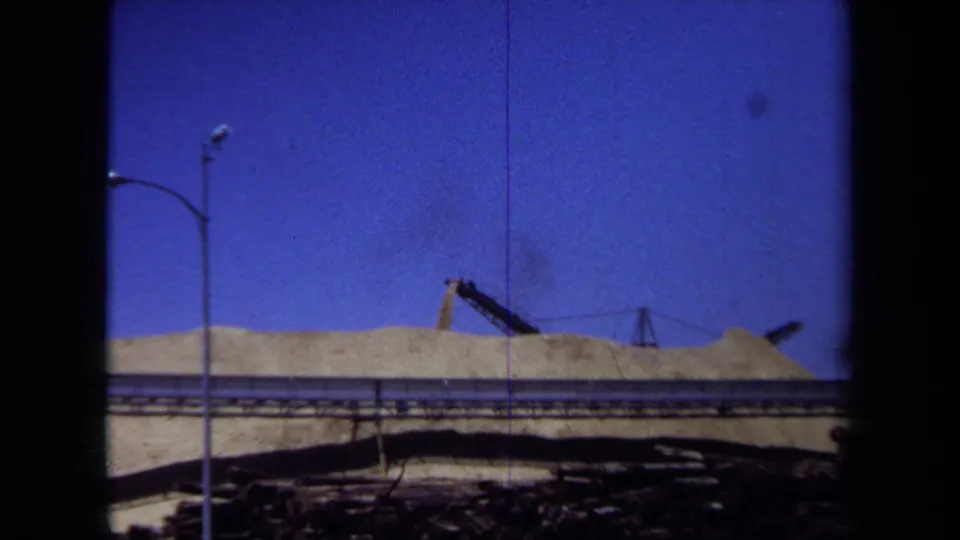 1971 Maine 8mm frame with a wide view of grain piles, conveyor machinery, foreground piping, and a streetlight at an industrial site.