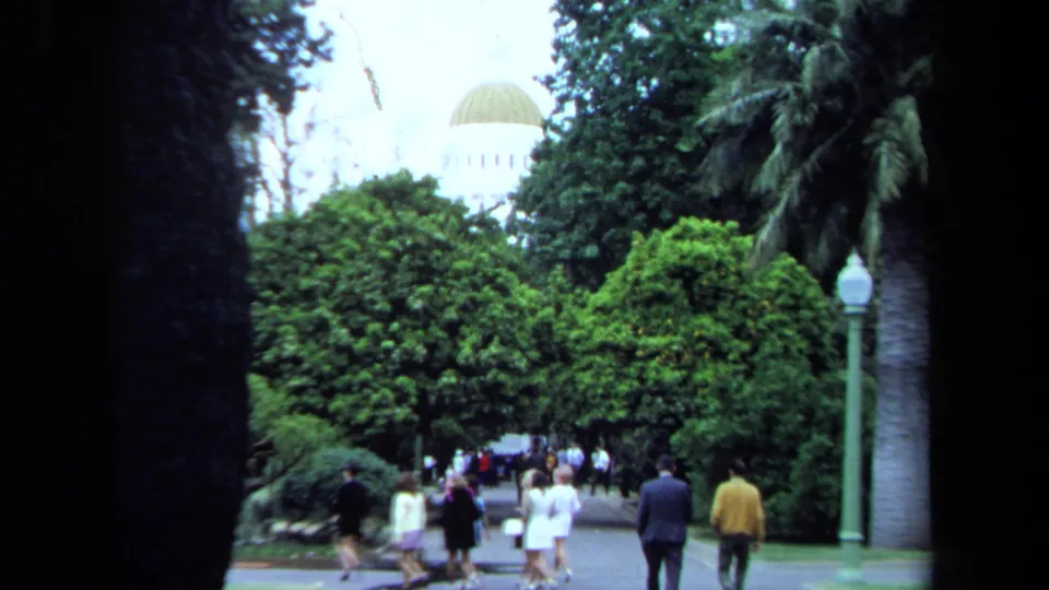 In footage identified as Maine in 1971, adults walk through a dense green park path with palm trunks, lampposts, and a pale domed building in the distance.