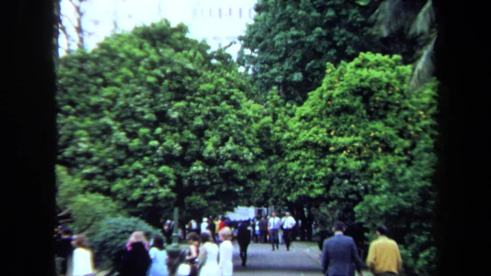A 1971 home-movie view shows adults walking away and toward the camera beneath thick green trees, with a pale building partly hidden above the foliage.
