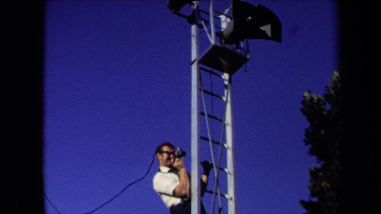 In Sacramento 1971 footage, a man in a white shirt and glasses stands on a tall ladder with a camera against a deep blue sky.