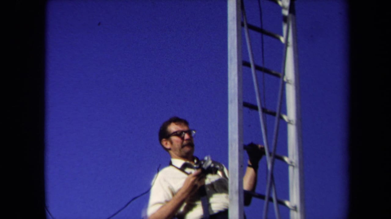Photographer with camera stands beside a tall ladder with blue sky behind him.