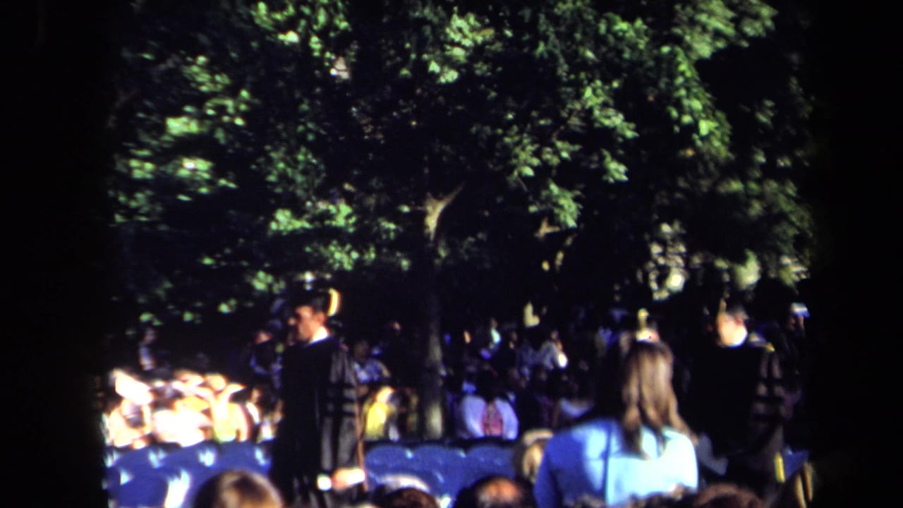 1971 Sacramento home-movie frame showing an outdoor night ceremony with spectators, blue chairs, and dark-clothed participants under trees.