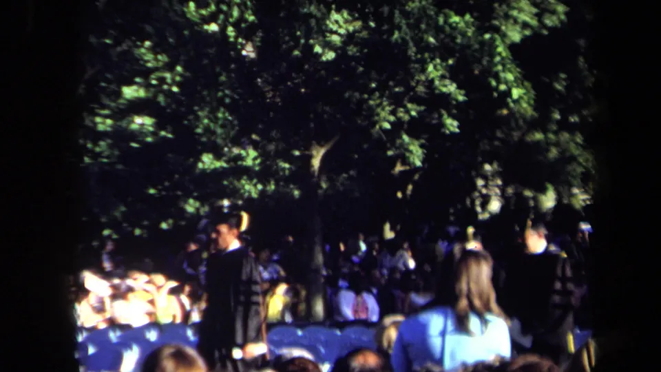 1971 Sacramento home-movie frame showing an outdoor night ceremony with spectators, blue chairs, and dark-clothed participants under trees.