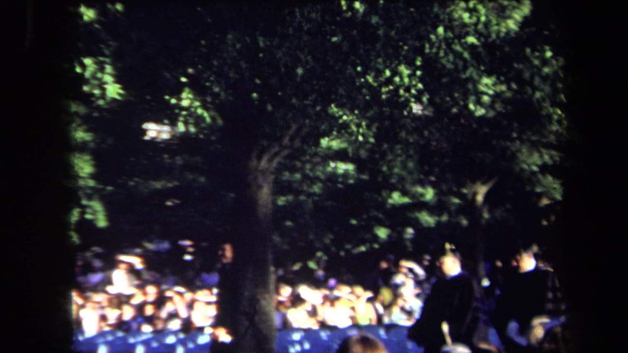 Outdoor Sacramento ceremony frame with a crowd, blue seating, and formal figures standing beneath floodlit tree cover.