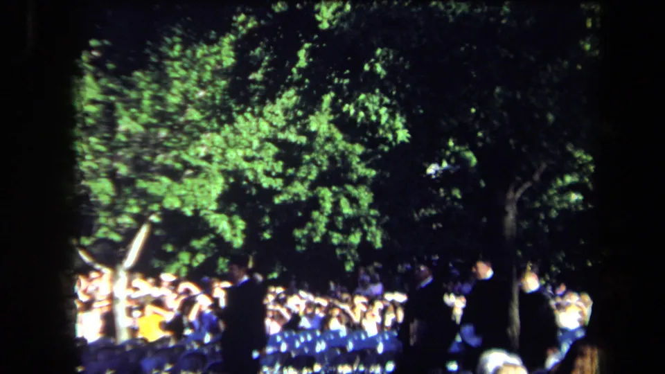 Home-movie frame from a 1971 Sacramento outdoor ceremony, with a crowd seated below trees and participants crossing the scene.