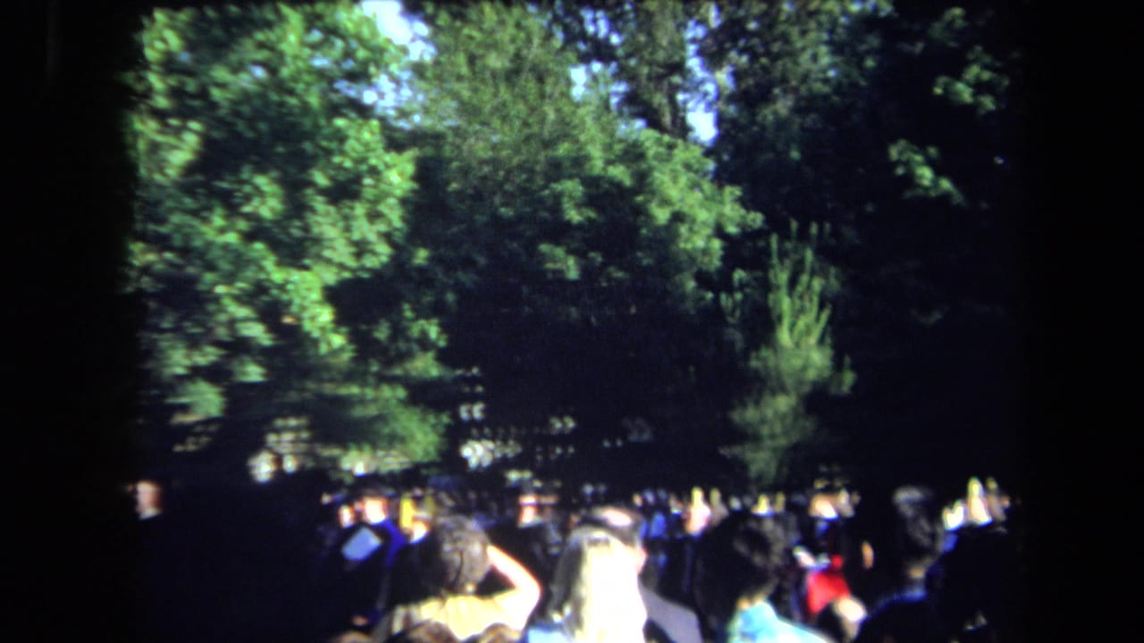 1971 Sacramento home-movie frame showing spectators and graduates gathered in a shaded park during an outdoor graduation event.