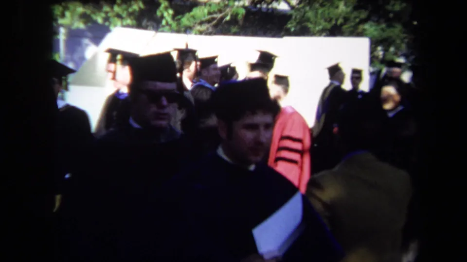 Close handheld 1971 Sacramento graduation view with mortarboards, dark gowns, a red robe, and spectators near a pale wall.