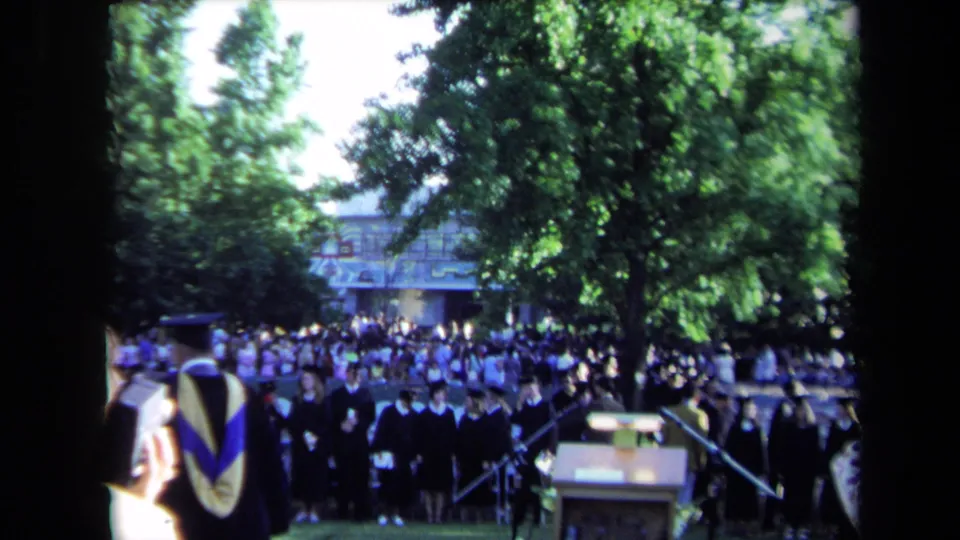 Wide frame of the Sacramento convocation showing a podium, robed participants, and a large outdoor audience under trees.