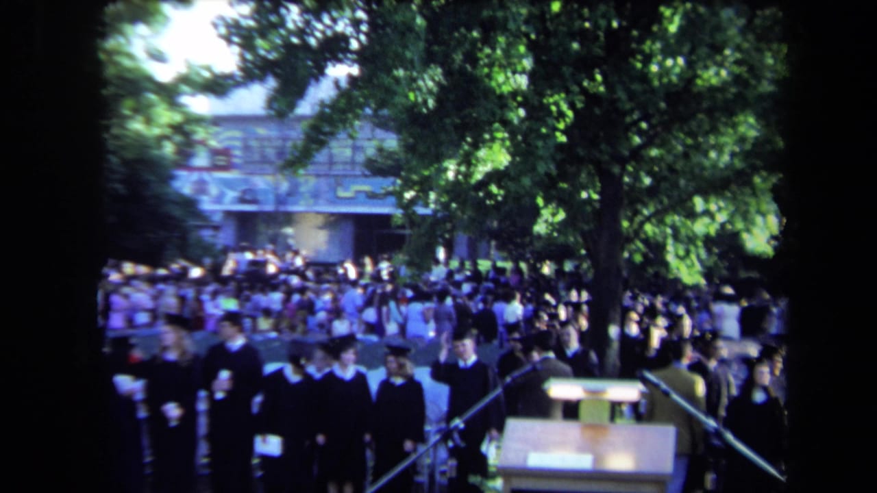 1971 Sacramento graduation scene with robed graduates assembling near microphones and a lectern under leafy trees.