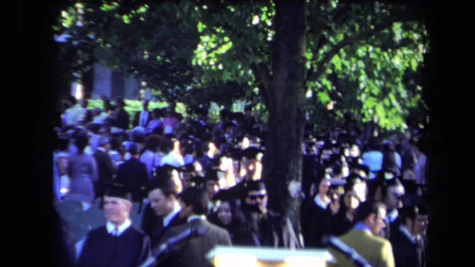 Shaded Sacramento graduation crowd in 1971, with robed students and spectators passing near a large tree and microphone stand.