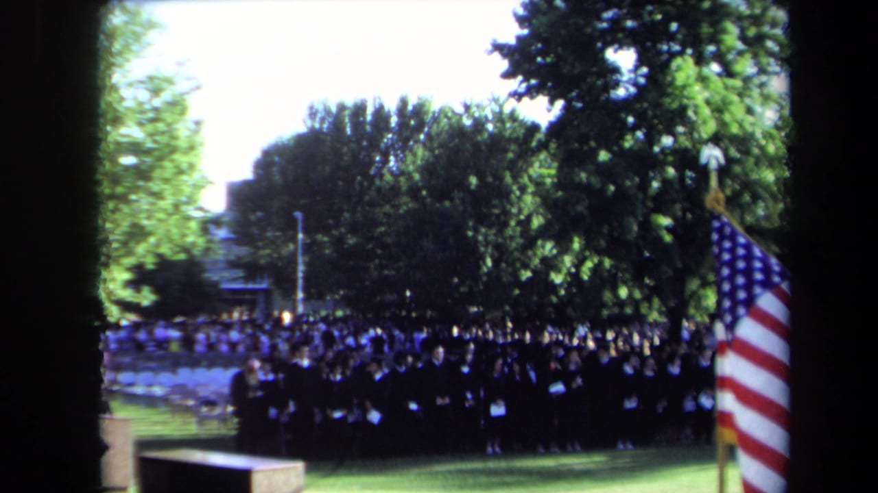 Wide Sacramento 1971 graduation view with rows of students in black caps and gowns near an American flag on a lawn.