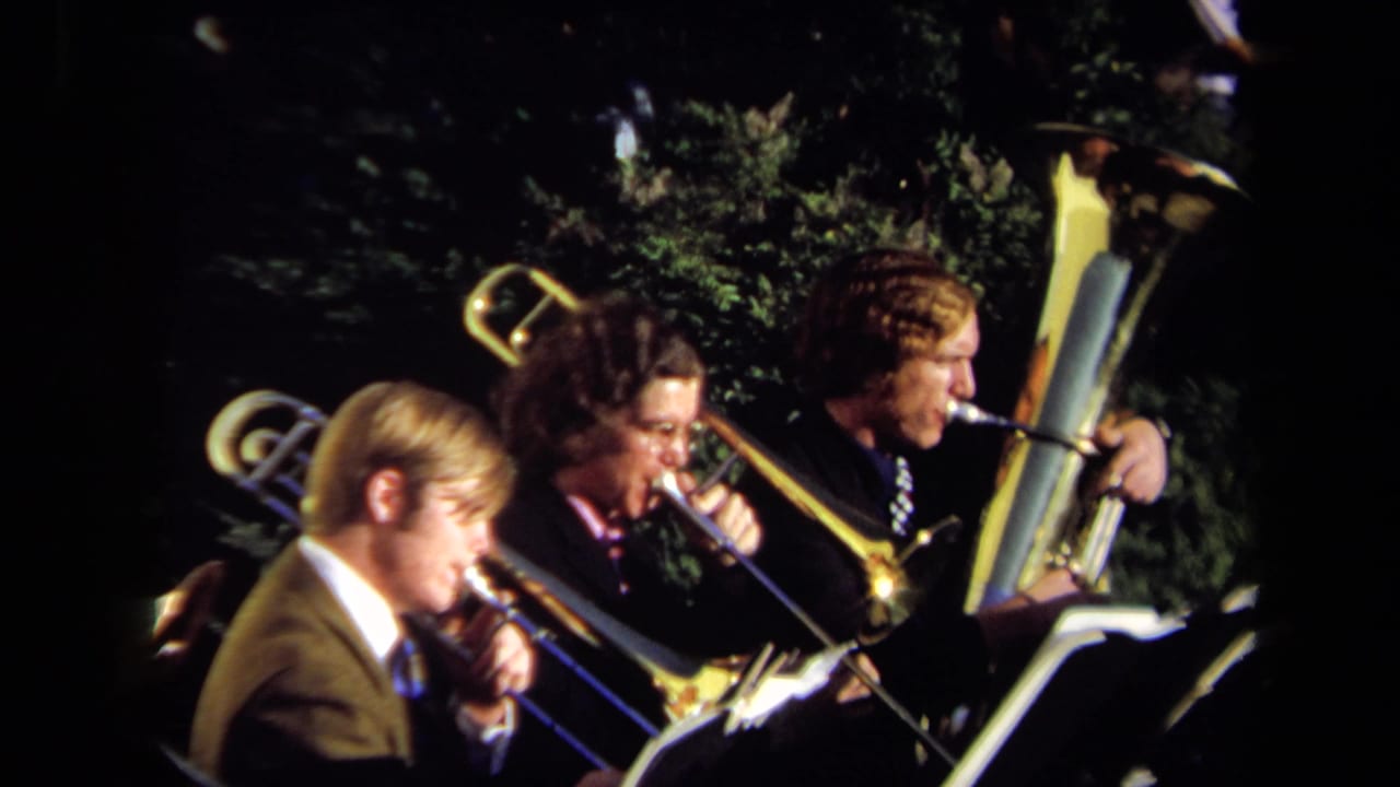 Three young men in suits play brass instruments beside music stands outdoors.