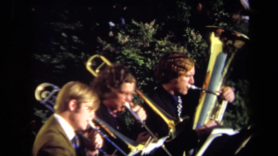 A close 8mm view shows three suited brass musicians performing outdoors in Sacramento, with stands and leafy background visible.